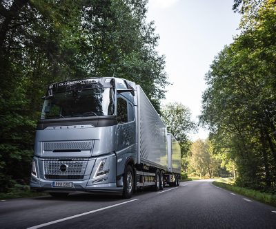 Volvo kamion sa prikolicom vozi se asfaltiranim putem kroz zeleni šumski pejzaž, snimljen iz niskog prednjeg ugla pri dnevnom svjetlu.