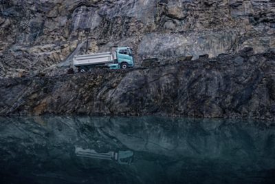 Elektryczna wywrotka Volvo Trucks jadąca wzdłuż skalistej półki kamieniołomu nad spokojną wodą.