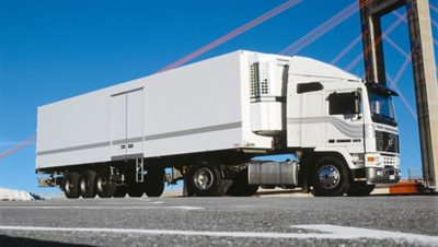 Weißer Volvo F12 Lkw mit Kühlanhänger, geparkt auf einer Brücke unter strahlend blauem Himmel und roten Tragseilen.