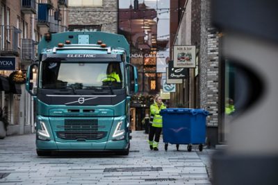 Elektryczny samochód ciężarowy Volvo stojący na wąskiej ulicy handlowej, podczas gdy operator przetacza duży niebieski pojemnik.