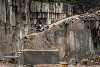 Samochód ciężarowy Volvo z przyczepą platformową ostrożnie jedzie wąską, gruntową drogą między stromymi ścianami skalnymi w kamieniołomie.