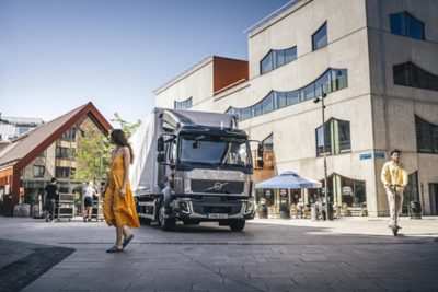 Samochód dostawczy Volvo porusza się po ruchliwym placu dla pieszych, obok spaceruje kobieta, a przed nowoczesnymi budynkami mężczyzna jedzie na hulajnodze elektrycznej.