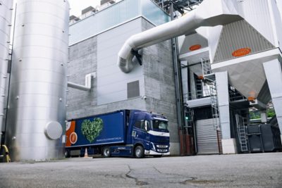 Volvo truck with a blue cab and a box trailer featuring a large graphic of a heart‑shaped foliage design is parked beside tall industrial buildings made of concrete and metal. The truck is positioned on a paved area near pipes, vents, and exterior structural elements of the facility.
