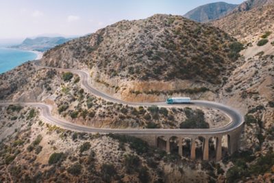 Aerial view of a Volvo truck with a box trailer travelling along a winding mountain road built into steep, rocky terrain, with the road curving sharply across multiple levels and coastal water visible in the distance.