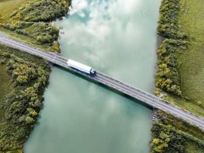 Nákladní automobil Volvo se skříňovým návěsem zachycený v pohledu shora přejíždí úzký most přes klidnou tyrkysově zbarvenou vodní hladinu se zelenými břehy řeky po obou stranách.