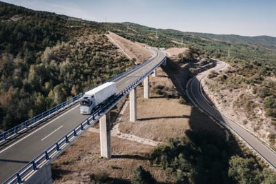 Samochód ciężarowy Volvo z naczepą skrzyniową jadący po podwyższonym moście autostradowym przez górzysty, zalesiony krajobraz.