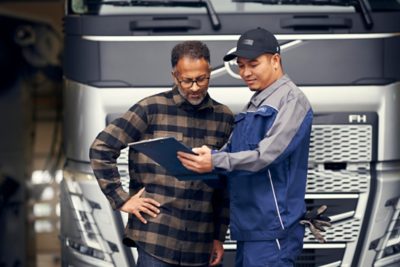 Two men reviewing documents on a clipboard in front of a Volvo truck, discussing service or maintenance details.