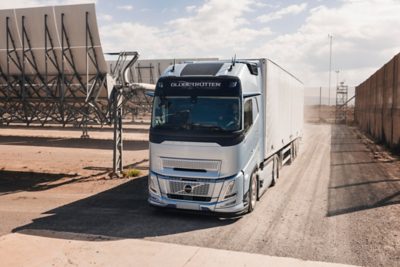 Nákladní automobil Volvo se skříňovým návěsem jede po prašné obslužné komunikaci vedle řad velkých solárních panelů v průmyslovém areálu.