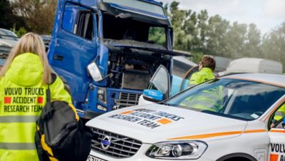 Das Unfallforschungsteam von Volvo Trucks untersucht den Unfallort mit einem beschädigten blauen Lkw und einem weißen Einsatzfahrzeug von Volvo.