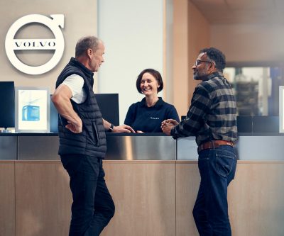 To kunder taler med en Volvo Trucks-repræsentant ved en receptionsskranke, mens et Volvo-logo kan ses i baggrunden.