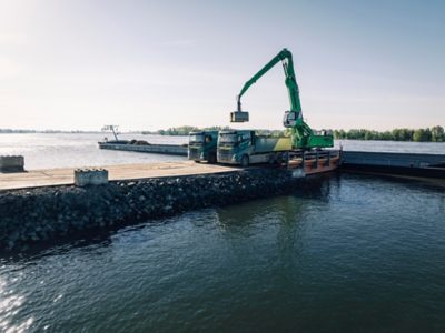Two Volvo trucks are positioned on a narrow embankment beside the water while a large green material‑handling excavator loads a pallet or container into the bed of one of the trucks. The scene takes place at a waterfront worksite with calm water and tree‑lined shoreline in the background under clear daylight.