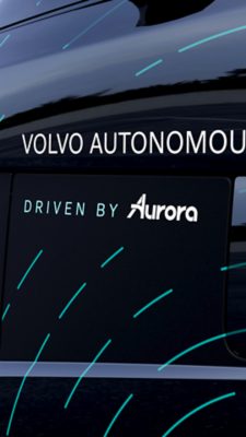 Autonomous Volvo semi-truck with the Aurora driver standing in front of a warehouse.