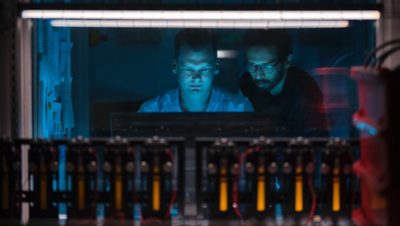 In the foreground, battery components are seen. In the background, two men are looking at a screen.