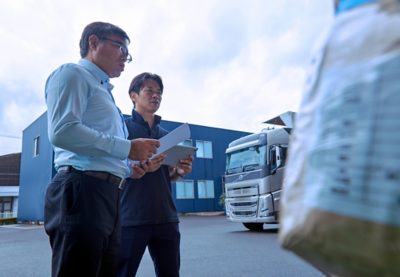 Two men stand outside near an industrial building, discussing paperwork and a tablet.