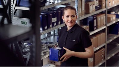 Inside a warehouse full of shelves. A woman carrying a box with a spare part from Volvo.