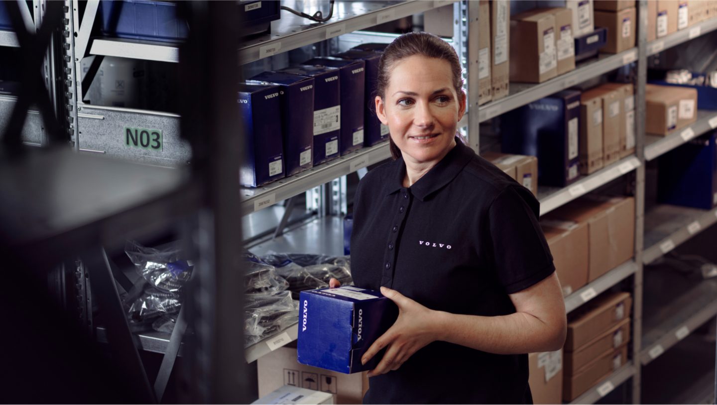 Inside a warehouse full of shelves. A woman carrying a box with a spare part from Volvo.