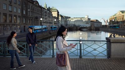A woman uses Volvo Buses bus fleet management system while crossing a bridge over a city canal. Two pedestrians in the periphery. 