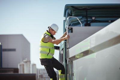 A person wearing high‑visibility safety clothing and a hard hat climbs the side steps of a Volvo truck, holding onto the grab handle while ascending. The scene takes place outdoors in an industrial environment with large structures visible in the background.