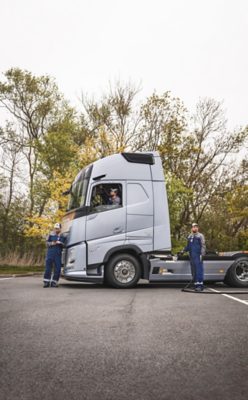 Darbuotojai, apsirengę mėlynais kombinezonais, dirba prie stovinčio sidabrinio „Volvo“ sunkvežimio ant asfalto: vienas sėdi kabinoje, kitas prijungia kabelį prie šono, trečias tikrina nešiojamąjį prietaisą, už jų – medžiai.